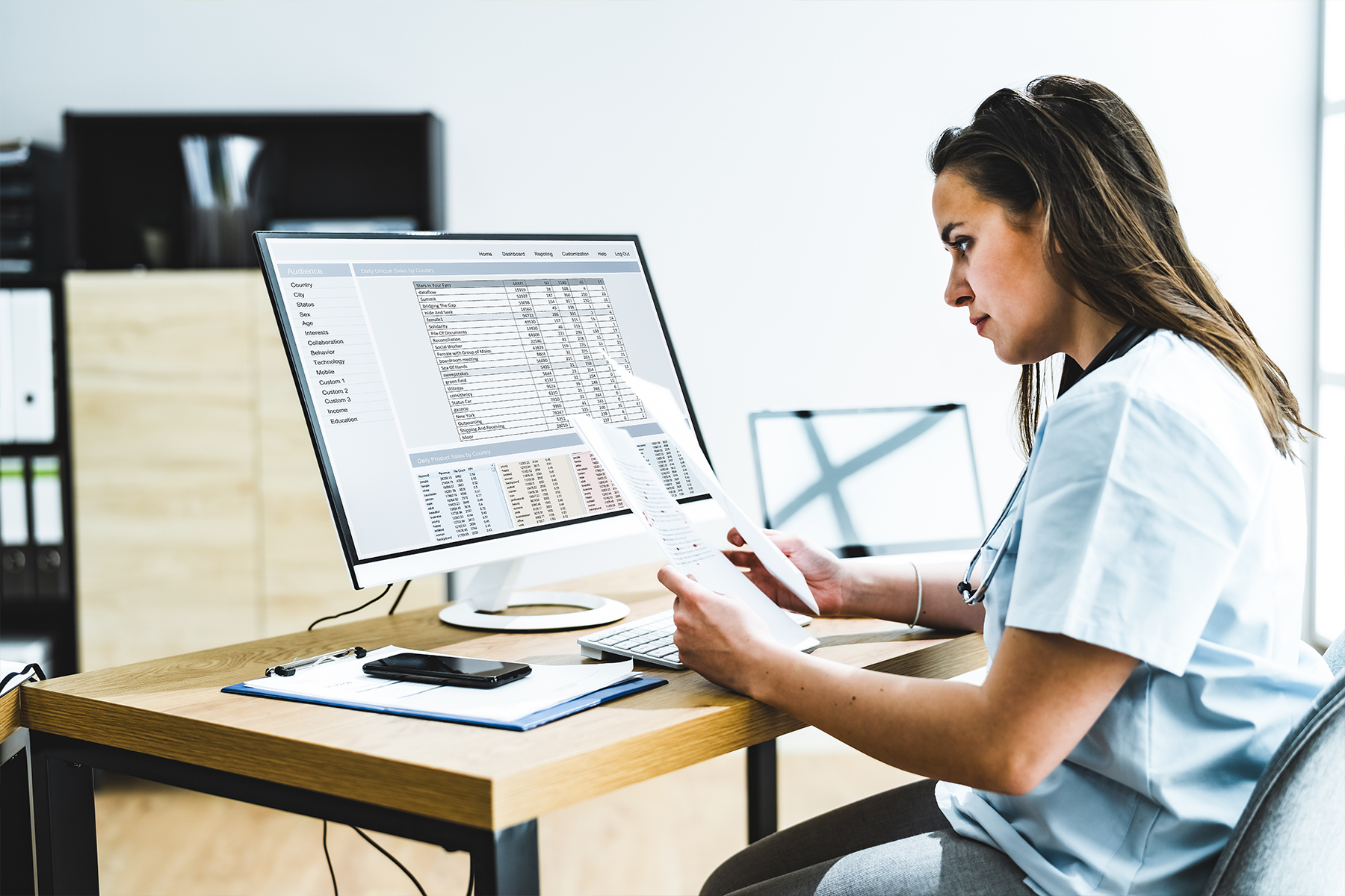 a woman in scrubs at a desk looking at paperwork with a spreadsheet on her computer