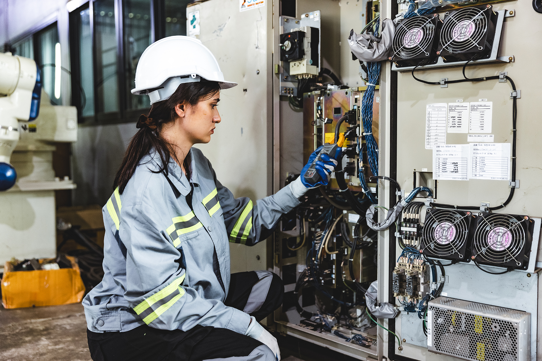 a young woman in a hardhat kneeling in front of a large electrical panel looking at a device in her hand