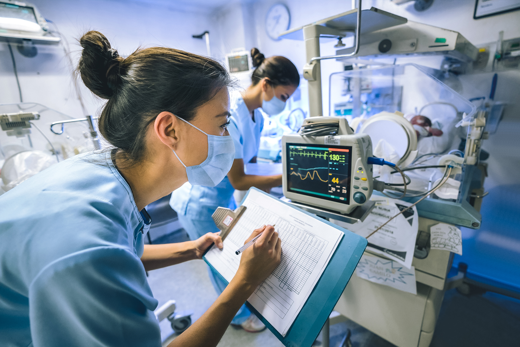 a woman in scrubs and wearing a facemask writes data on a chart while looking at a health monitor in the NICU